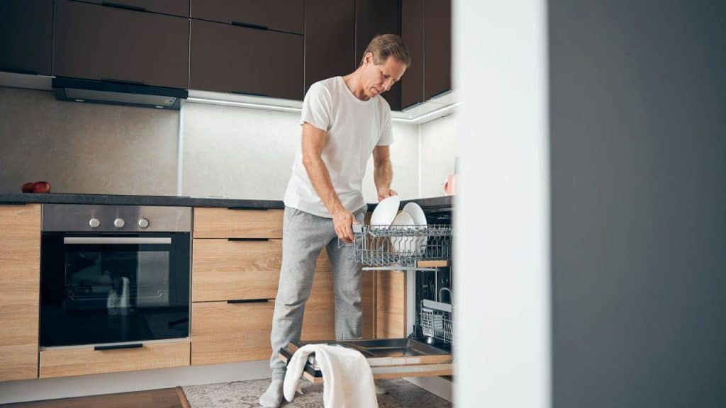 A man in a white shirt loads dishes into a dishwasher in a modern kitchen.