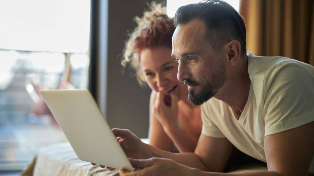 Man and woman lying on a bed together, smiling and looking at a laptop screen.