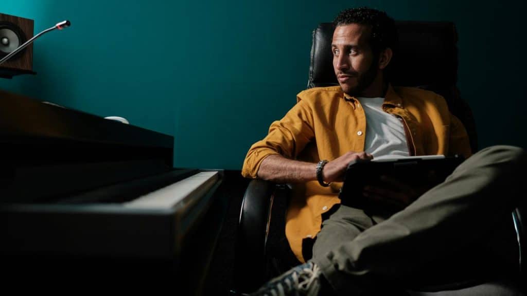 A man in a yellow shirt sitting in a chair holding a tablet near a piano.