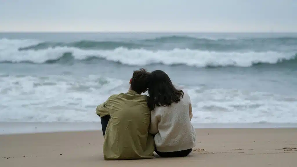 A couple sits side-by-side on the sand, looking out at the ocean waves under an overcast sky.
