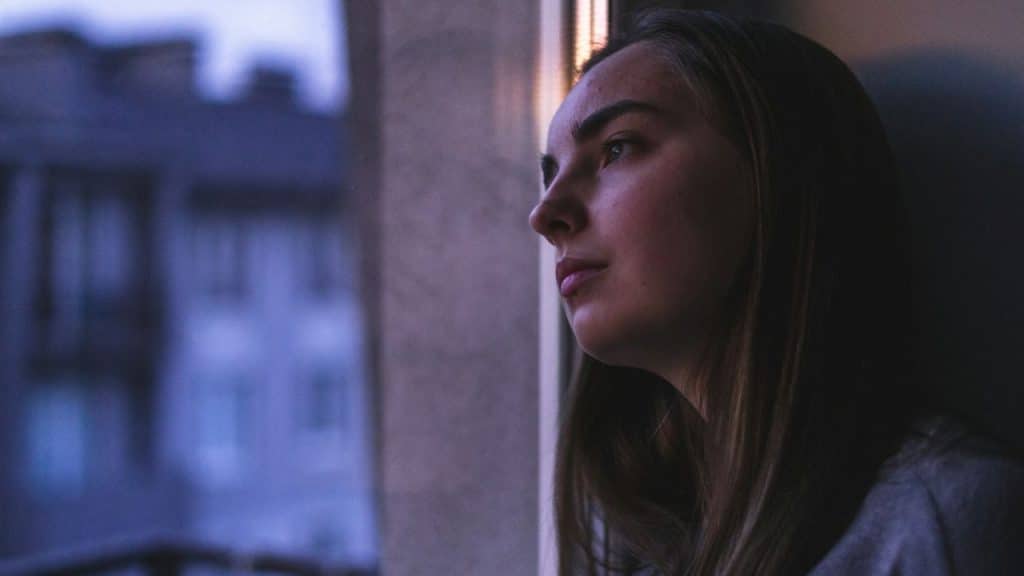 Woman with long hair looking out a dark window at a blurry, blue city building.