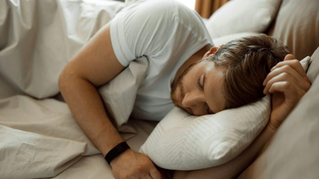 A man with light brown hair is sleeping in bed, tucked under white covers.