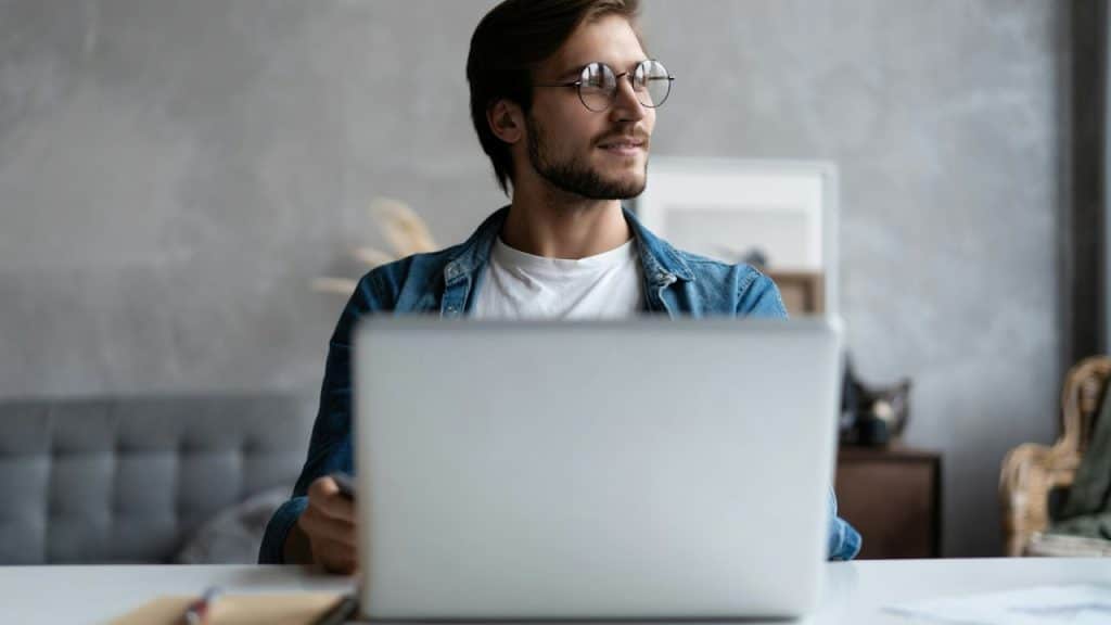 A smiling man in a denim shirt and glasses looks away from a laptop at a bright desk.