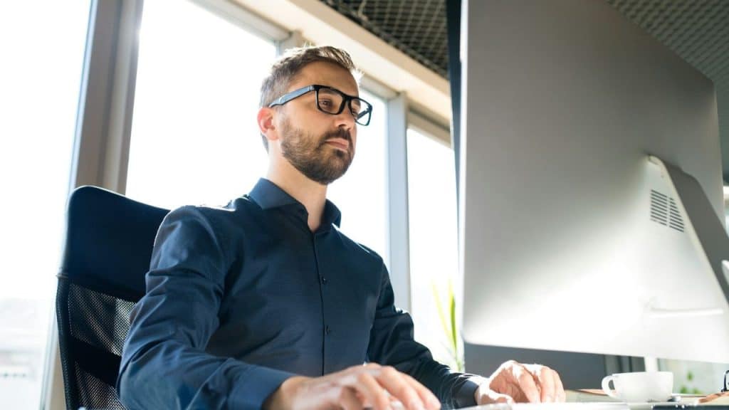 A man with a beard and dark-rimmed glasses is intently working on a computer in a bright office.