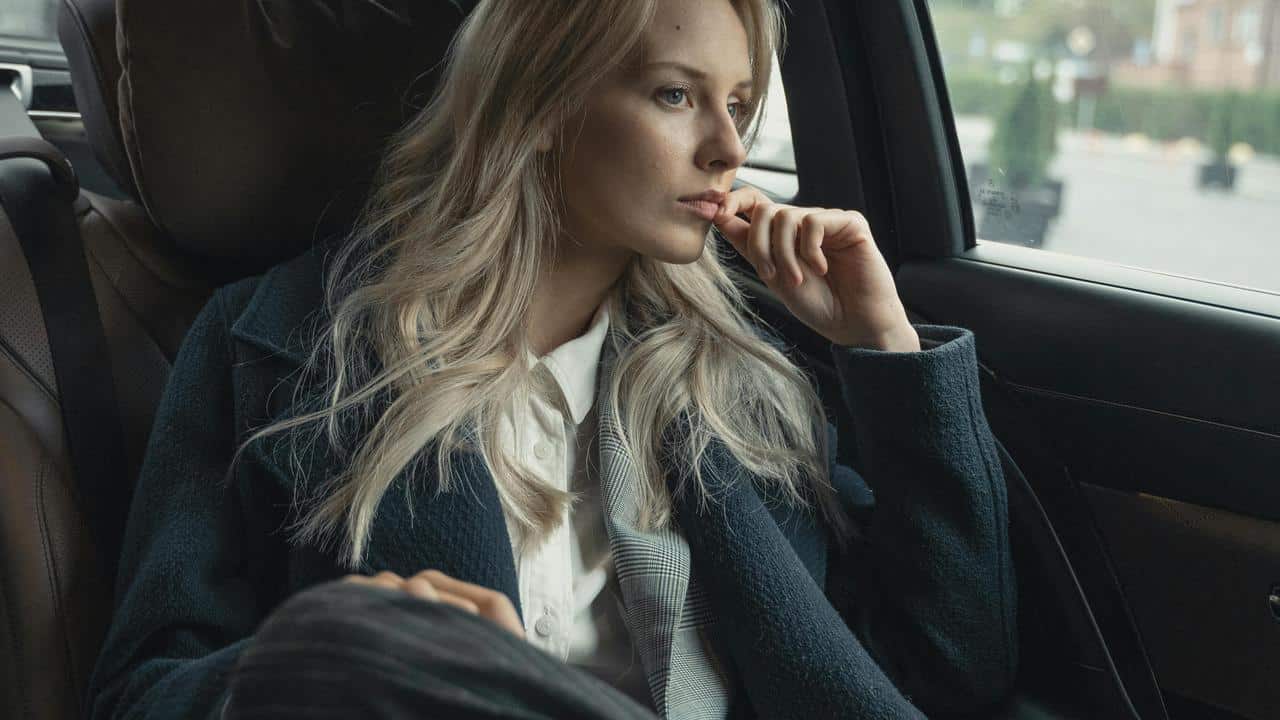 A person with long blonde hair sitting in a car and looking out the window thoughtfully.