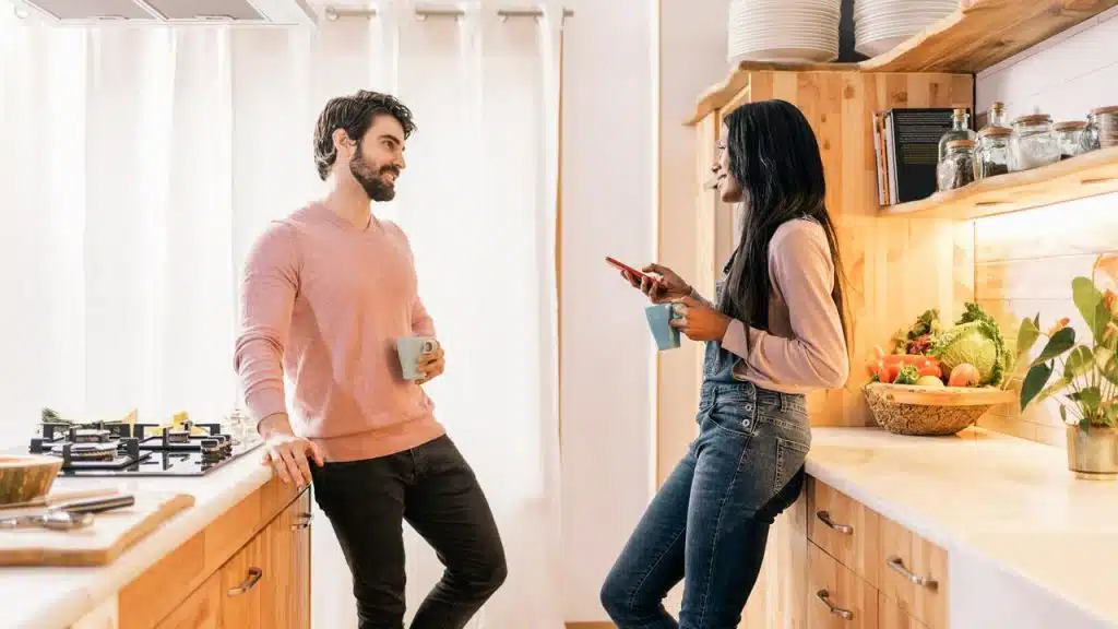 A man and a woman standing in a kitchen, talking and holding mugs.