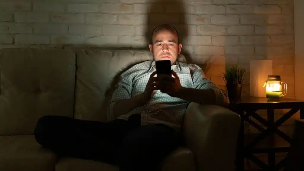 A man sits on a couch in the dark, looking down at his phone's glowing screen.