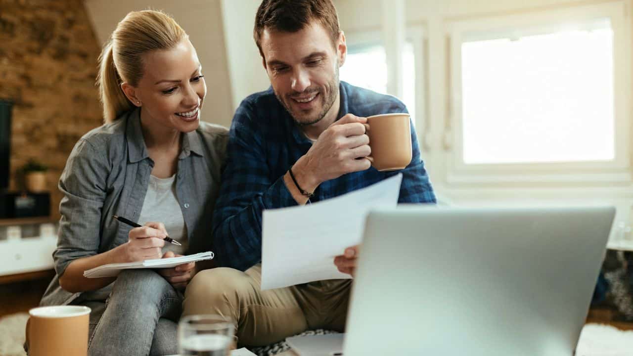 A couple reviews documents and a laptop, with the woman taking notes and the man holding a mug.