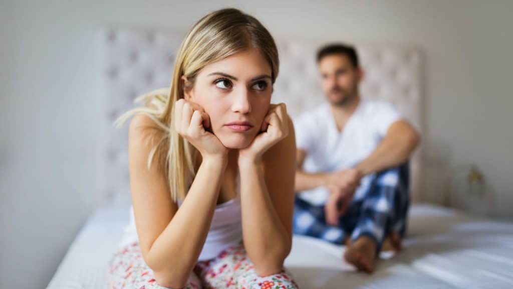 A blonde woman looking sad sits on a bed with a man blurred in the background.