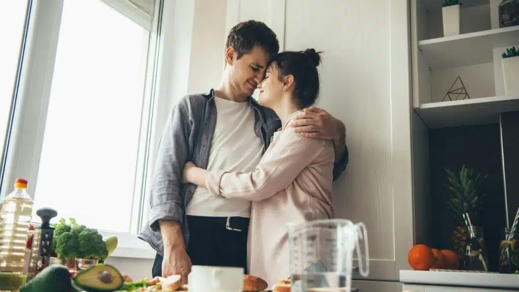 Smiling couple standing close, embracing in a brightly lit kitchen with food on the counter.