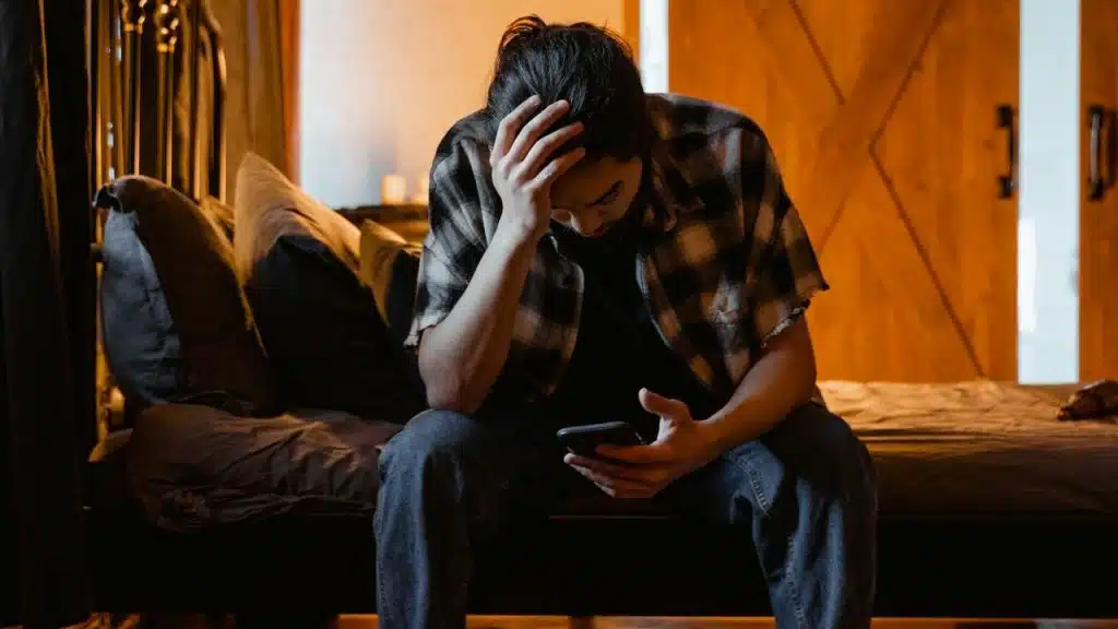 Man sitting on a bed, looking distressed while holding a phone.