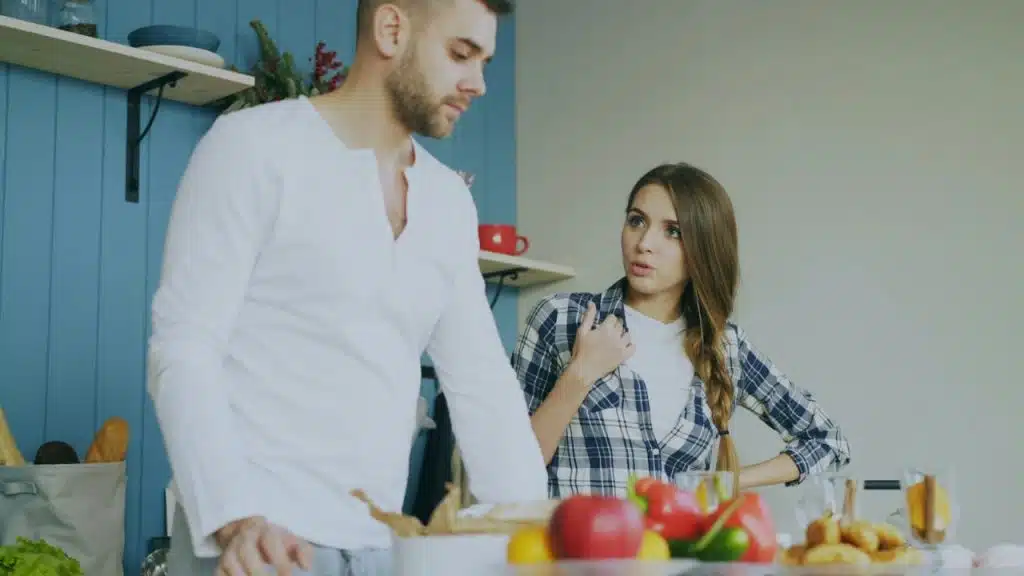 A woman with a braid talks to a man in a white shirt in a kitchen with vegetables on the counter.