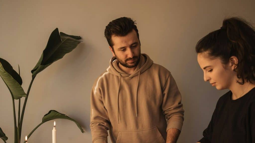 A man in a tan hoodie is standing next to a woman and a potted plant.