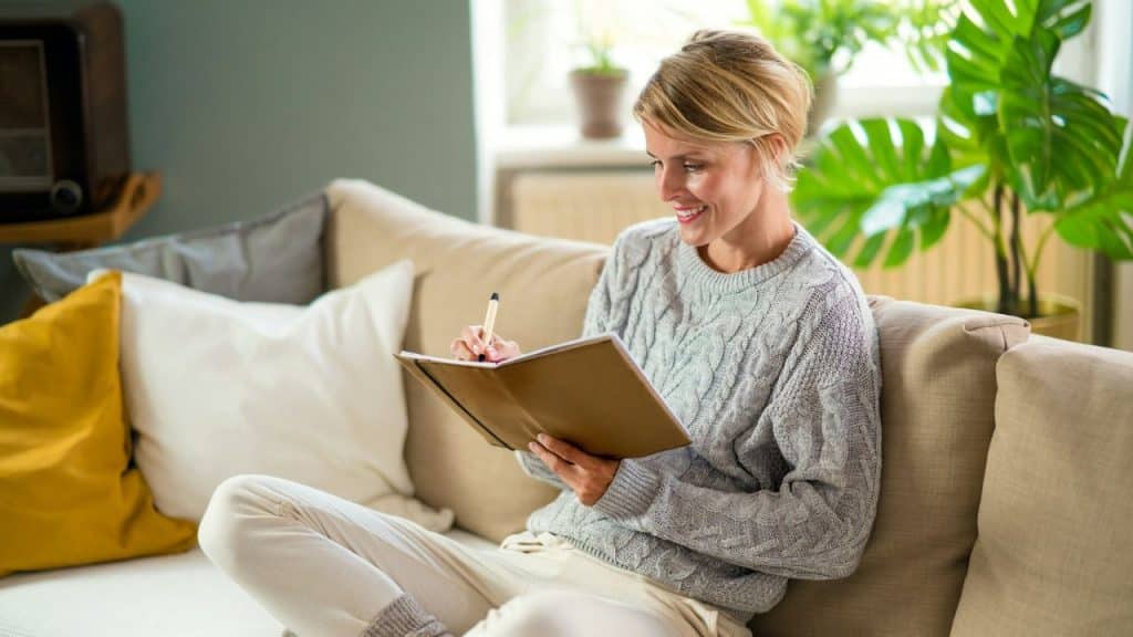 A smiling woman in a gray sweater writes in a journal on a light-colored couch.