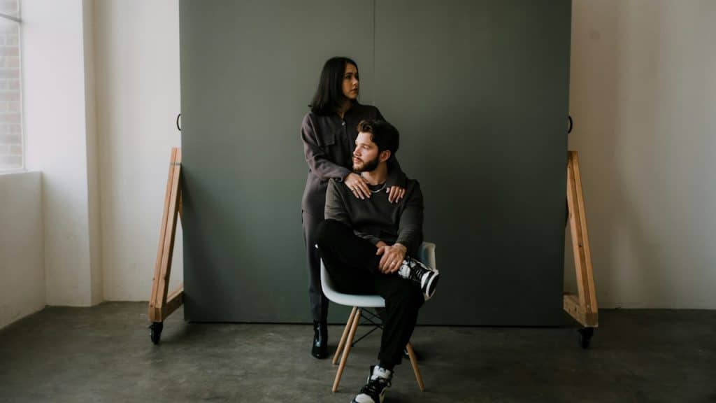 A man in a black jacket is sitting on a white chair while a woman is standing behind him.