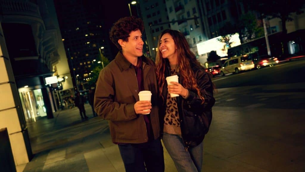 A happy couple holding coffee cups walks on a city street at night.