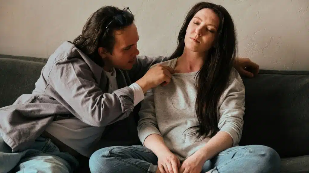 A man leans in closely, touching a woman's shoulder, as she sits with closed eyes.