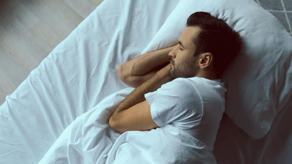 Man with a beard and white shirt sleeping on a white bed with arms behind his head.