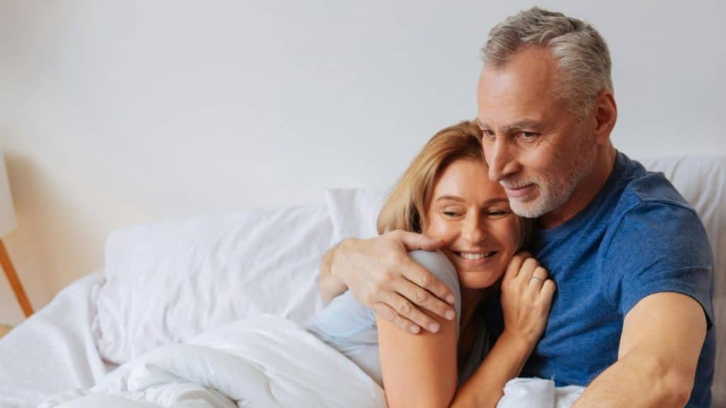 Man with beard and graying hair embracing a smiling woman in a bed.