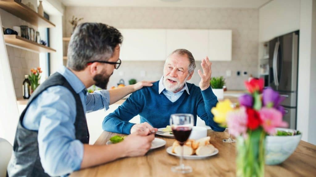 Middle-aged man in blue sweater and glasses talking to younger man at dinner table.