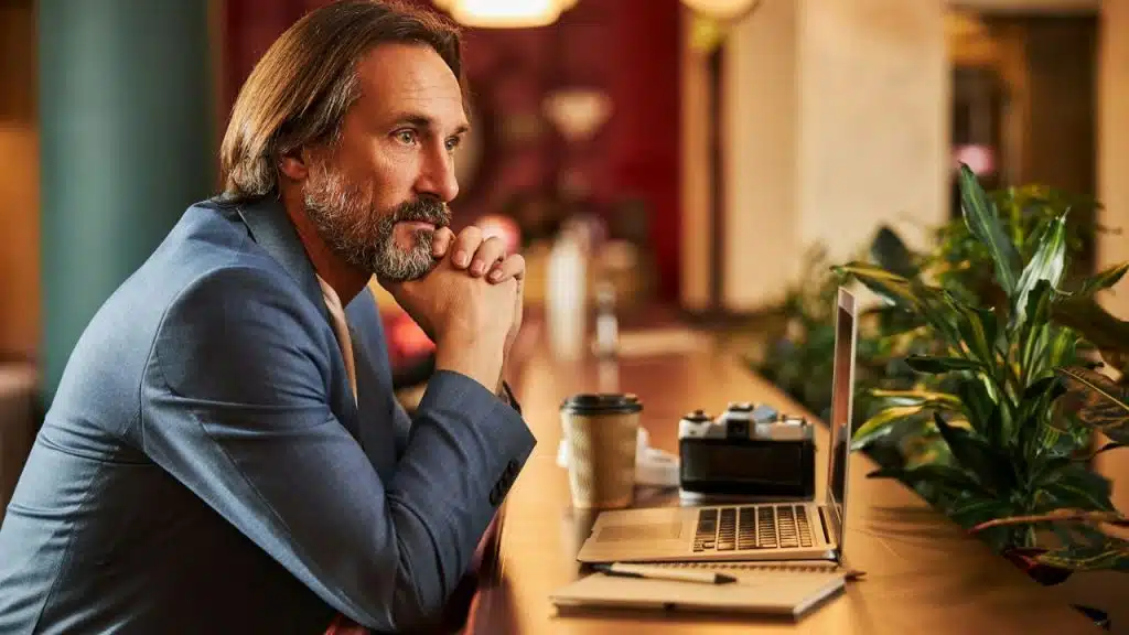Older man in blue suit jacket looks thoughtfully at a laptop on a counter.