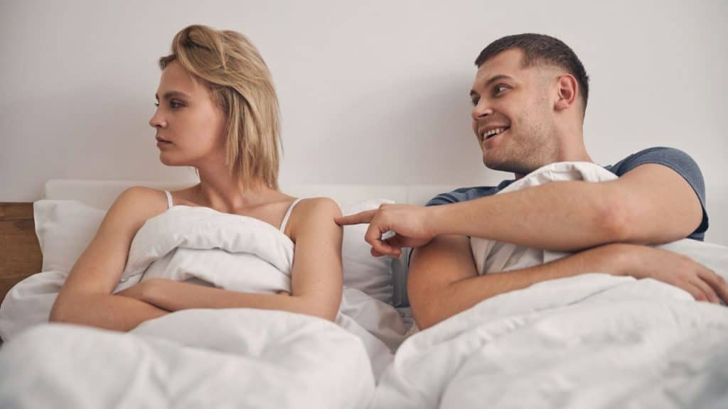 A smiling man in bed pokes a woman who is lying next to him with her back turned and arms crossed.