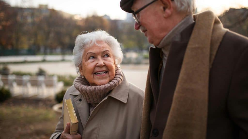 An elderly couple walking outdoors, smiling at each other, with the woman holding a book.