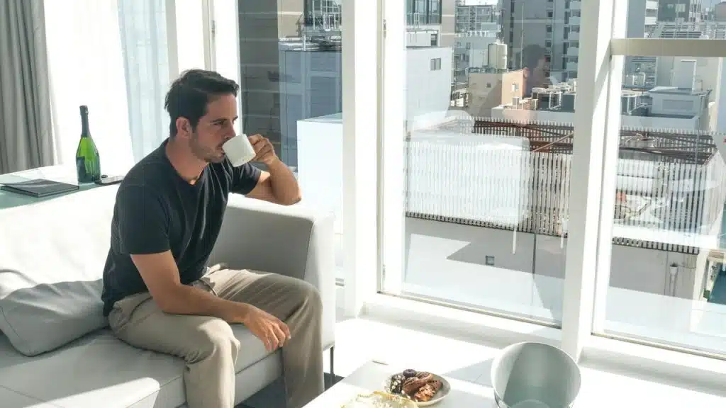 A man sitting on a couch drinking coffee by a large window.