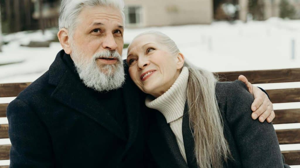 A senior couple sitting on a bench outdoors in winter, embracing each other warmly.