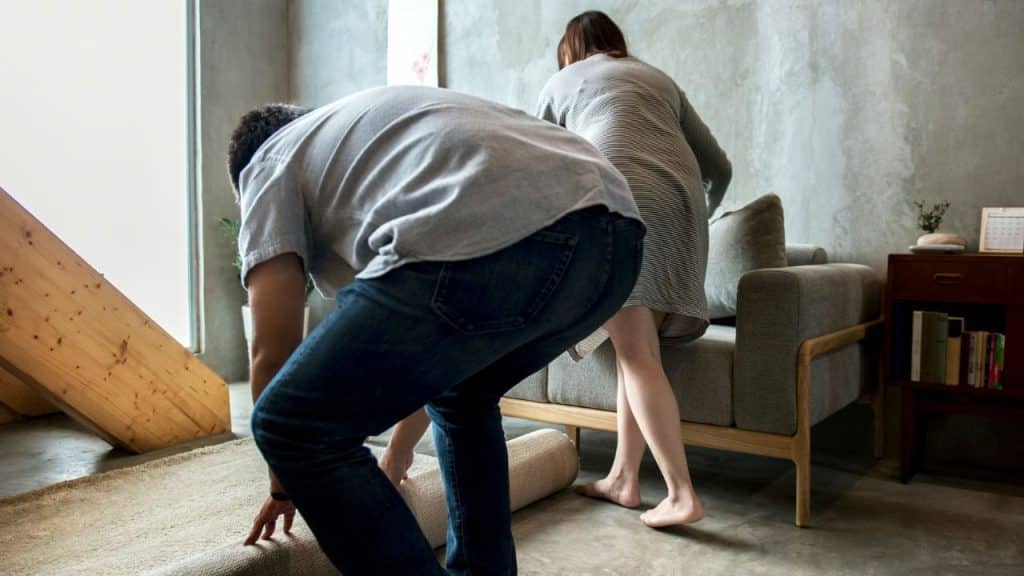 A man and woman work together to roll up a rug in a modern living room.