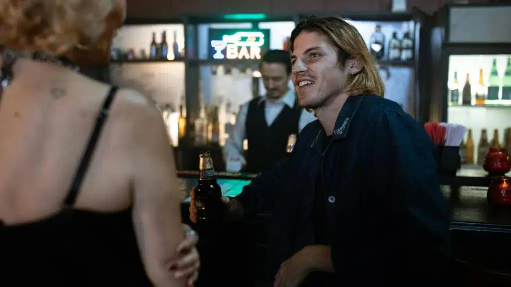 A man sitting at a bar talking to a woman while holding a beer.