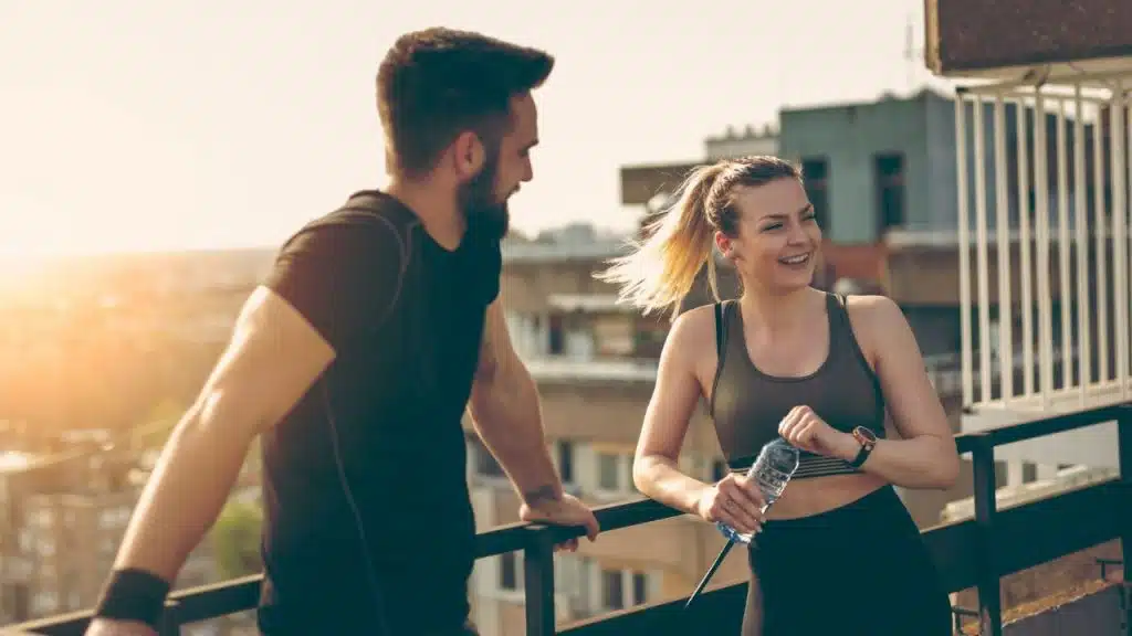 A man and woman in athletic wear talk on a rooftop while holding a water bottle.