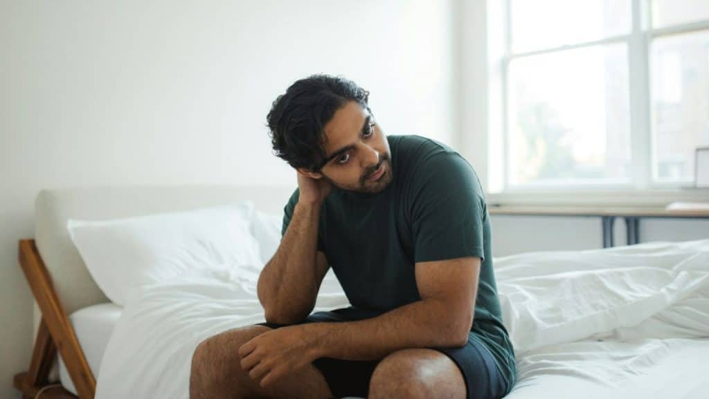 A man in a green t-shirt sits on a bed, looking over his shoulder.