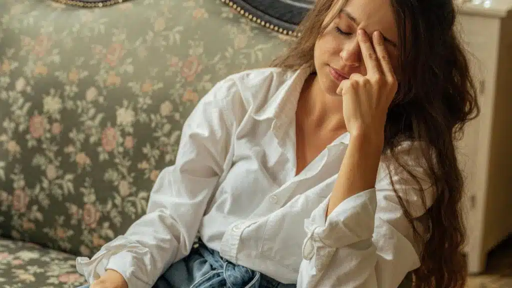 A woman resting on a vintage sofa with her eyes closed and hand on her forehead in a sunlit room.