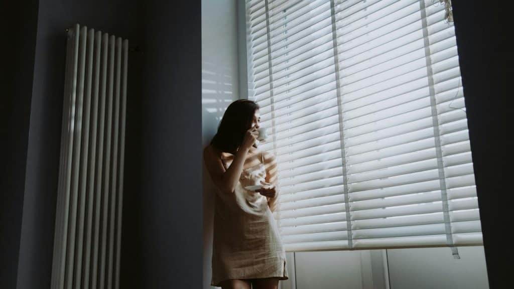 A woman standing by a window with blinds, holding a cup and looking outside.