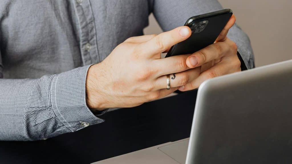 A person holding a smartphone sits at a laptop.