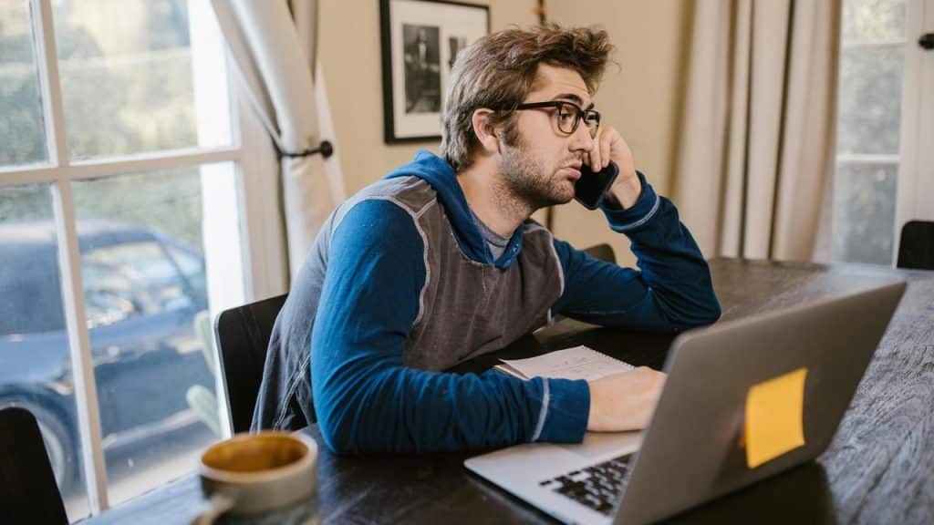 A man sitting at a table working on a laptop while talking on a phone.