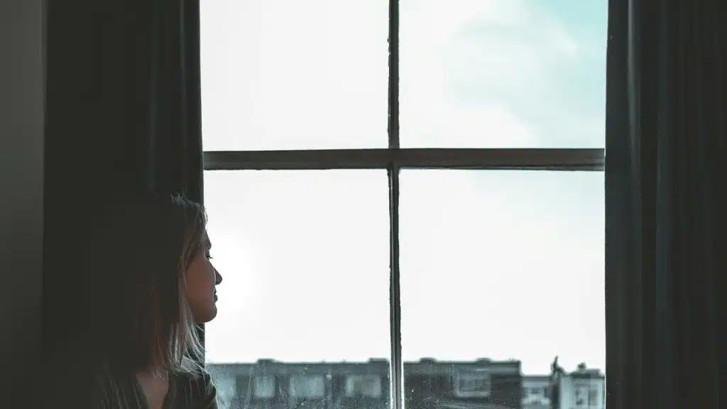A woman in profile gazing out a large window with curtains and city buildings beyond.