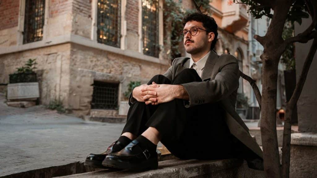 Man in a blazer and glasses sitting on an outdoor stone ledge looking thoughtful.