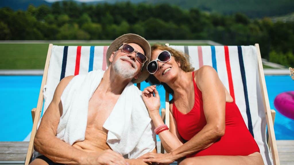 A cheerful couple sitting by a swimming pool outdoors in the backyard looking at the camera.