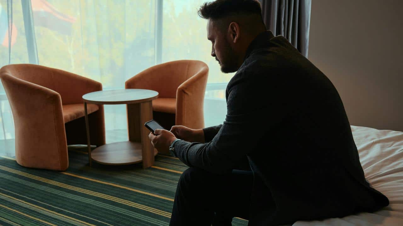A man in a dark suit sitting on a bed and looking at his phone in a hotel room.