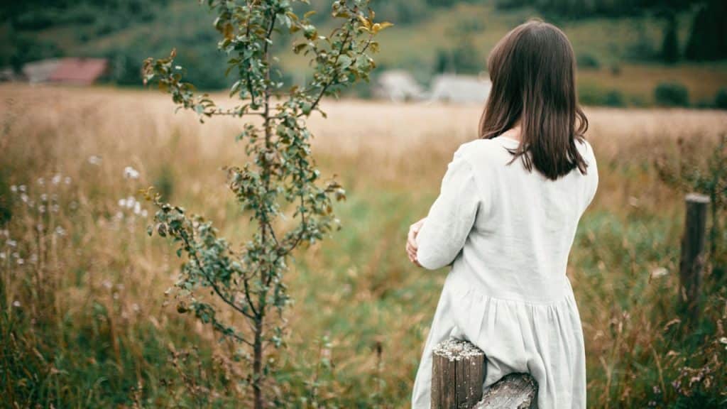 A stylish girl in linen dress standing at an aged wooden fence among herbs and wildflowers looking at the field.