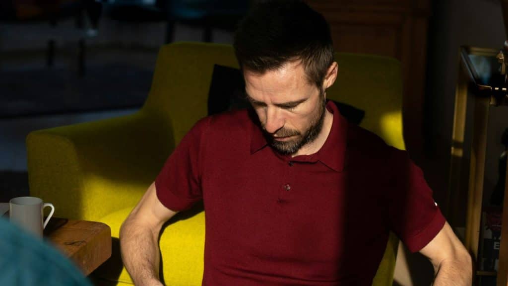 Man in a red polo shirt sitting in a brightly lit yellow armchair, looking down.