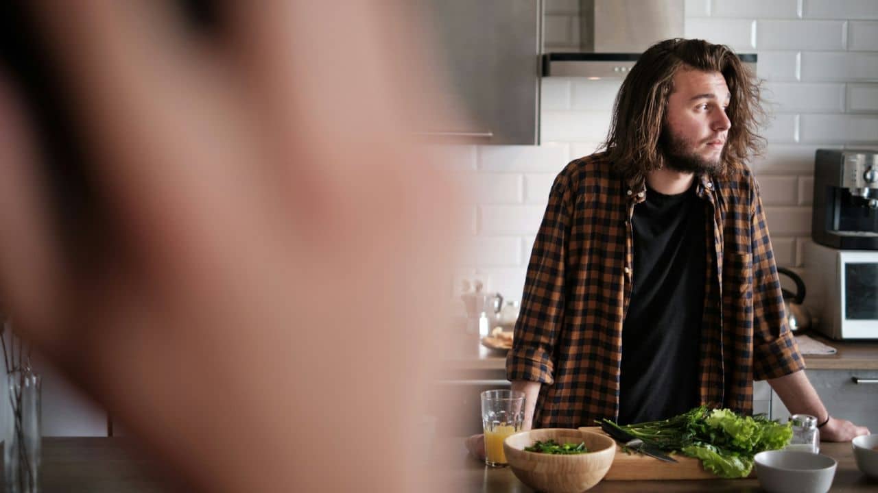 Man with long hair and a plaid shirt standing in a kitchen with greens on the counter.