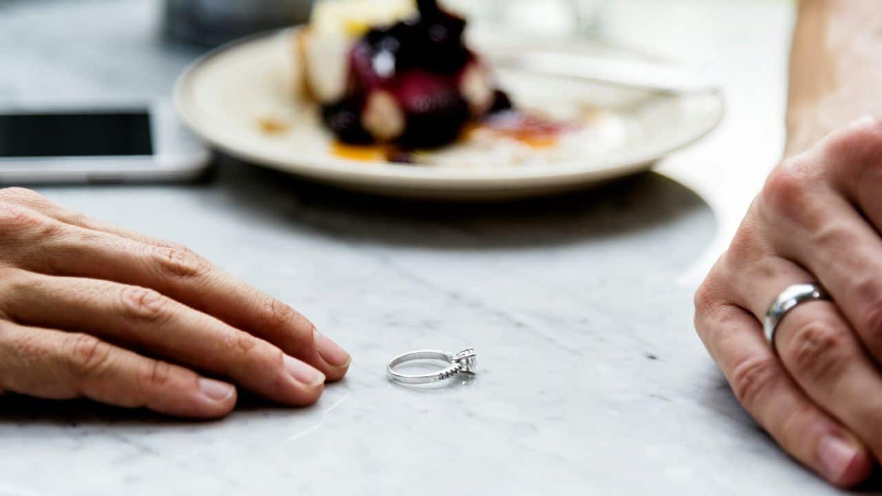 Close-up of a diamond engagement ring resting on a marble table between two hands.