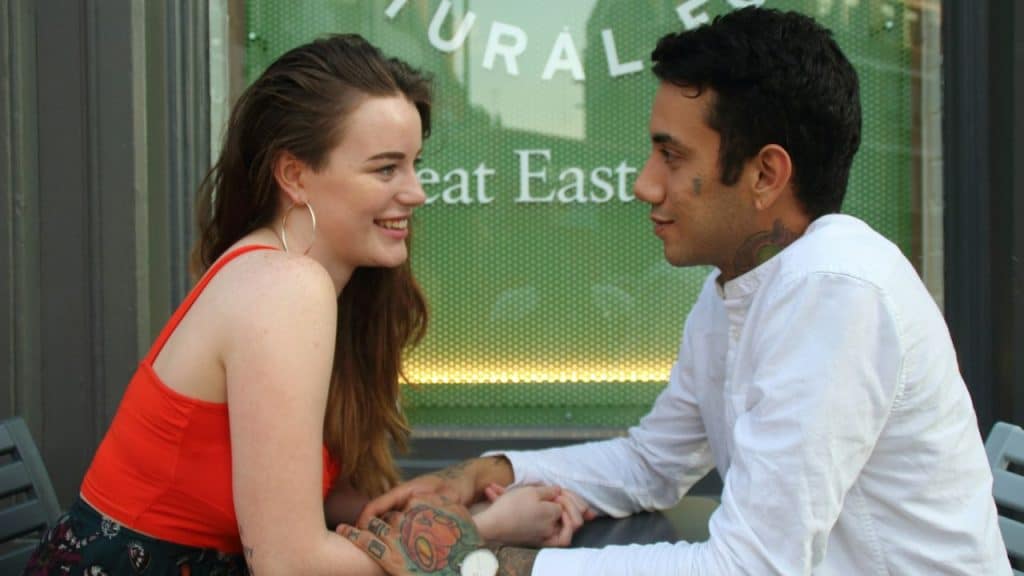 Smiling woman and man with tattoos sitting across a table, holding hands.
