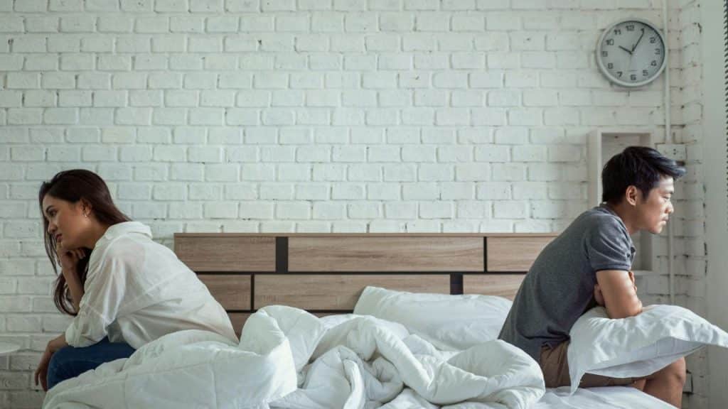 Man and woman sit turned away from each other on a bed with a white brick wall.