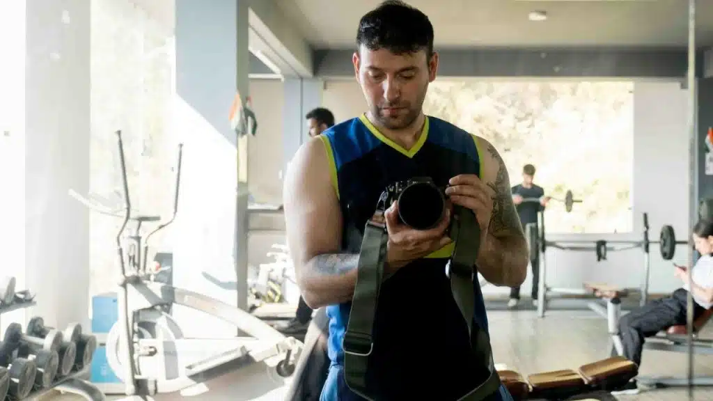 Man in a tank top holding a camera, seemingly taking a picture in a gym.