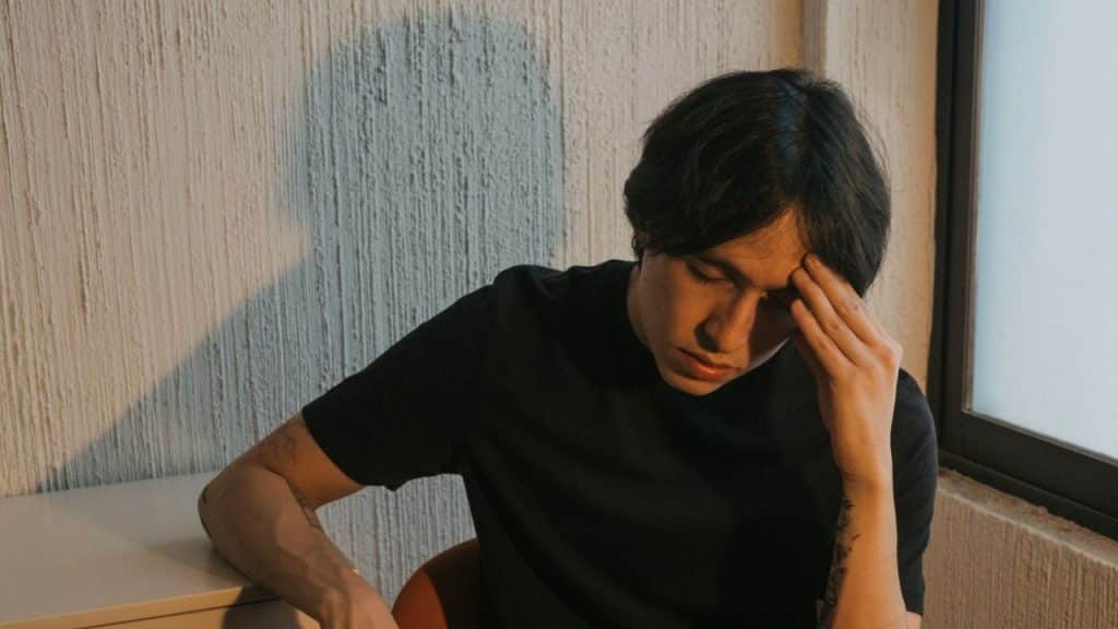 A young man in a black T-shirt sits, holding his forehead with one hand.