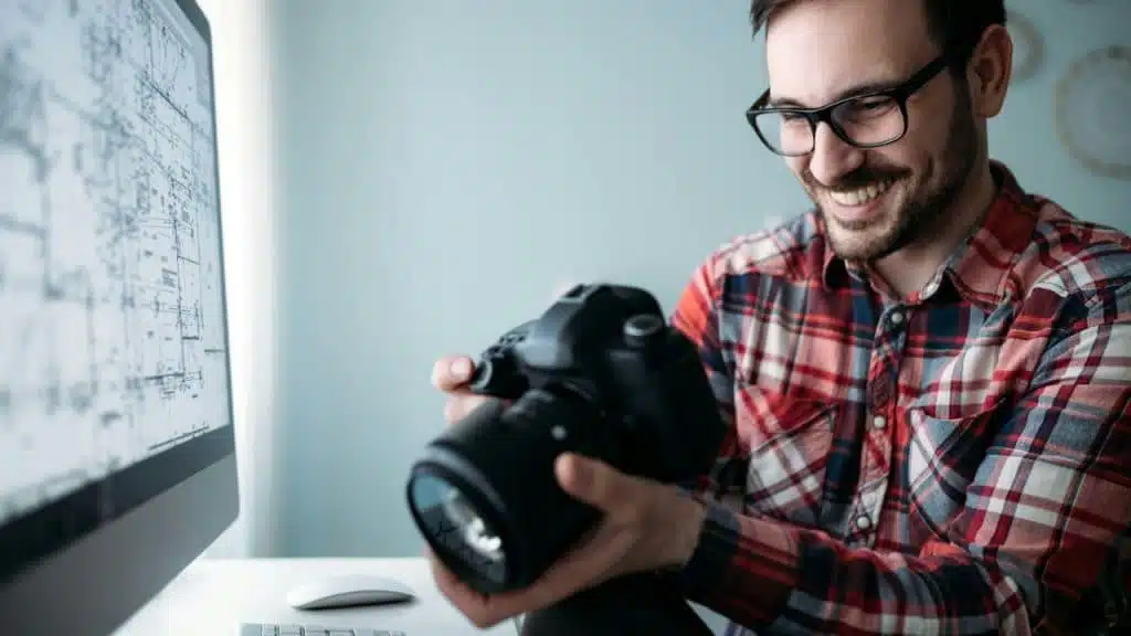 Smiling man in glasses and a plaid shirt holds a professional camera near a computer screen.